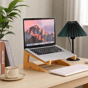 May include: A silver laptop computer rests on a light-colored wooden stand. The screen displays a mountain landscape. Nearby are a white ceramic mug on a saucer, a white wireless keyboard, and a dark green pleated lamp on a black base.