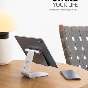 May include: A silver metal tablet stand holding a gray tablet, with a smartphone resting nearby. The text STAND YOUR LIFE and An excellent addition to your desk. is visible at the top. The stand features a circular cutout and a cylindrical hinge.
