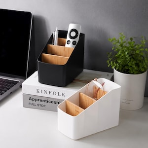 May include: Two desk organizers with matte black and white plastic shells and light brown bamboo dividers. Each organizer features three tiered compartments for storage. The black unit holds a pen and remote, while the white unit holds glasses.