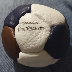 May include: A spherical ball constructed from stitched panels of white, dark blue, and brown leather. The white leather panel features the text Johannes Von Rechtel written in black ink. The surface of the leather has a visible textured grain.