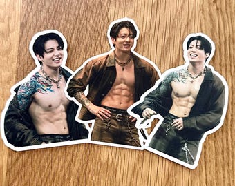 May include: Three die-cut stickers featuring a person with dark hair, visible tattoos on the arms, and a muscular torso. The person wears an open brown jacket, dark pants with a visible waistband, and a silver chain necklace in each image.