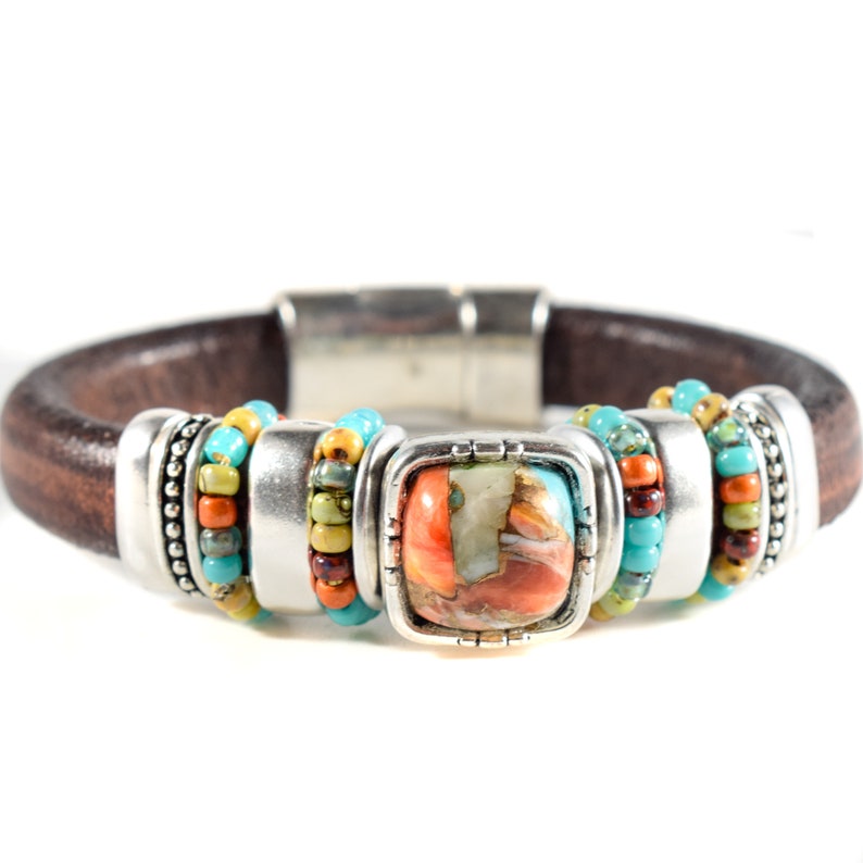 May include: Brown leather bracelet with a square, multi-colored stone centerpiece. The bracelet features silver-toned metal accents and is adorned with turquoise, orange, and yellow beads. The bracelet has a magnetic clasp.