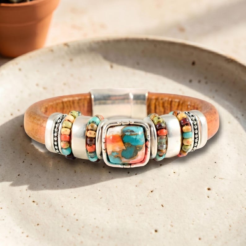 May include: A brown leather bracelet with a silver clasp and decorative silver accents. The bracelet features a square turquoise stone with orange and red hues, and colorful beaded sections.