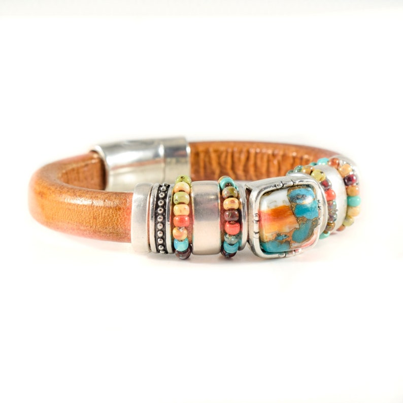 May include: A brown leather bracelet with silver-tone metal accents and a turquoise and orange stone. The bracelet features colorful beads in shades of red, orange, green, and blue. The bracelet has a magnetic clasp.