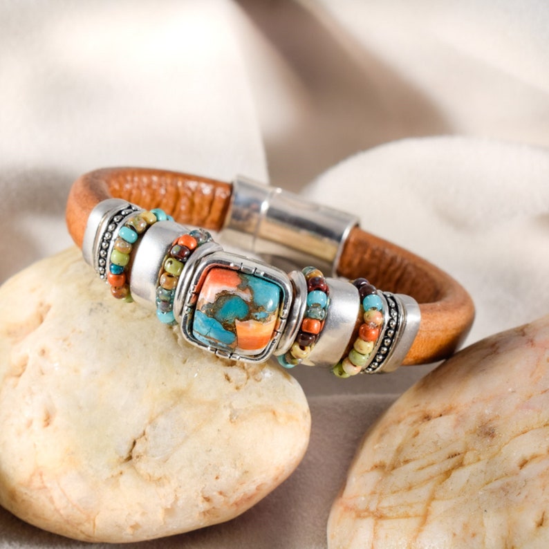 May include: A brown leather bracelet with a silver magnetic clasp. The bracelet features a square turquoise and orange stone centerpiece, surrounded by silver accents and colorful beaded details. The bracelet is resting on a light-colored stone.