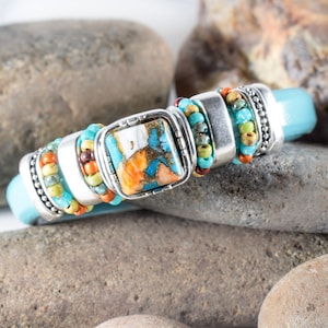 May include: A turquoise leather bracelet with silver-toned metal accents and a square, patterned stone. The bracelet features colorful beads in shades of turquoise, orange, and yellow. The stone has a unique pattern with shades of blue, orange, and white.