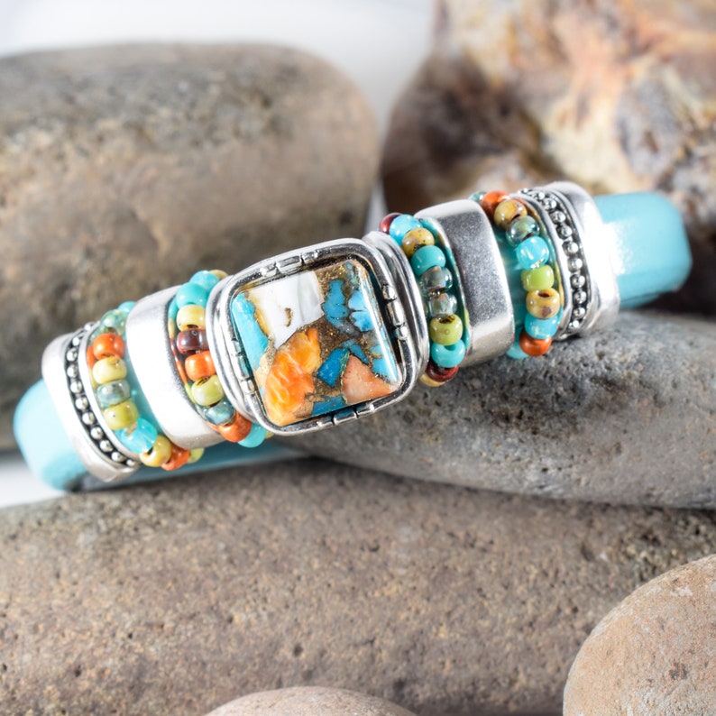 May include: A turquoise leather bracelet with silver-toned metal accents and a square, patterned stone. The bracelet features colorful beads in shades of turquoise, orange, and yellow. The stone has a unique pattern with shades of blue, orange, and white.