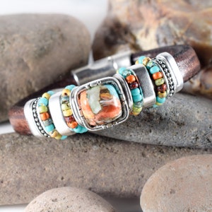May include: A brown leather bracelet with a silver magnetic clasp. The bracelet features a square, multi-colored stone set in a silver frame, surrounded by turquoise, orange, and yellow beads. The bracelet is approximately 0.75 inches wide.