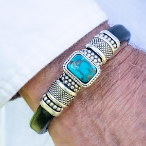 May include: A black leather bracelet with a rectangular turquoise stone centerpiece. The bracelet features silver-toned metal accents with a textured design. The bracelet is worn on a person's wrist.