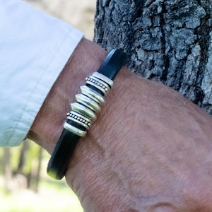 May include: Black leather bracelet with silver-colored metal accents. The bracelet features a series of stacked, ridged elements and smaller beaded sections. The bracelet is worn on a wrist, with a white shirt sleeve visible in the background.