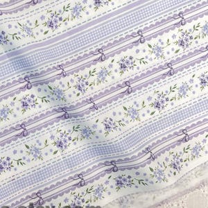 Könnte beinhalten: Weißer Stoff mit einem sich wiederholenden Muster aus lavendelfarbenen Blumen, Schleifen und Streifen. Der Stoff hat ein gewelltes Kantendesign und der Text "fabricmade.etsy.com" ist sichtbar. Eine große weiße Rose befindet sich in der oberen linken Ecke.