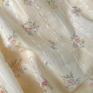 May include: A cream-coloured fabric with a floral pattern. The material has vertical stripes and is decorated with clusters of pink, yellow, and purple flowers. The texture appears soft and slightly textured, suggesting a delicate material.