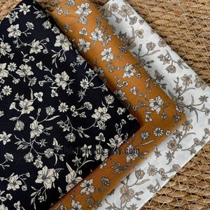 以下が含まれることがあります： 花柄の生地見本が3つ。1つは白花柄の黒、もう1つは白花柄のマスタードイエロー、3つ目は茶色の花柄の白。織り込まれた表面に生地が配置されています。