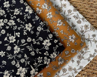 Pode incluir: Três amostras de tecido com padrões florais. Uma é preta com flores brancas, outra é amarelo mostarda com flores brancas e a terceira é branca com flores castanhas. Os tecidos estão dispostos sobre uma superfície tecida.