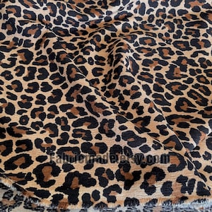 Könnte beinhalten: Nahaufnahme eines Stoffes mit Leopardenmuster. Der Stoff weist ein sich wiederholendes Muster aus schwarzen und braunen Flecken auf beigem Hintergrund auf. Der Stoff ist gefaltet und zeigt die Textur und die Musterabweichungen. Die Website-Adresse fabricmadeetsy.com ist sichtbar.