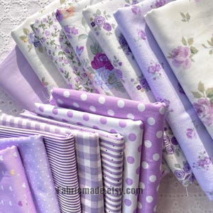 Puede incluir: Una colección de telas plegadas en varios tonos de morado y blanco. Los patrones incluyen flores, lunares, rayas y cuadros. Las telas están dispuestas en una exhibición en capas, mostrando los diferentes diseños y texturas.