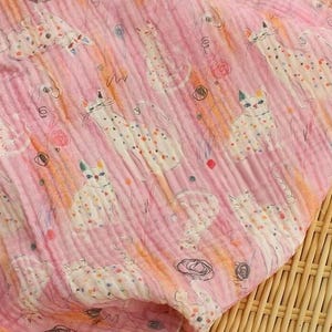 May include: A soft, crinkled pink fabric featuring a repeating pattern of white cats with colorful polka dots and small black circular scribbles. The textile is draped over a round, woven bamboo tray, showcasing a lightweight, layered texture.