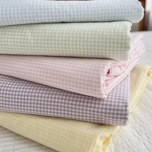 May include: A stack of folded bedding in pastel colors. The sheets are light blue, green, pink, purple, and yellow, all with a small gingham check pattern. The bedding is neatly stacked on top of each other.