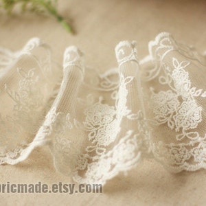 May include: Close-up of ivory lace trim with a floral design. The delicate fabric is gathered, creating a ruffled effect. The lace features a scalloped edge and is suitable for various sewing and craft projects.