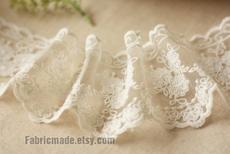 May include: Close-up of ivory lace trim with a floral design. The delicate fabric is gathered, creating a ruffled effect. The lace features a scalloped edge and is suitable for various sewing and craft projects.