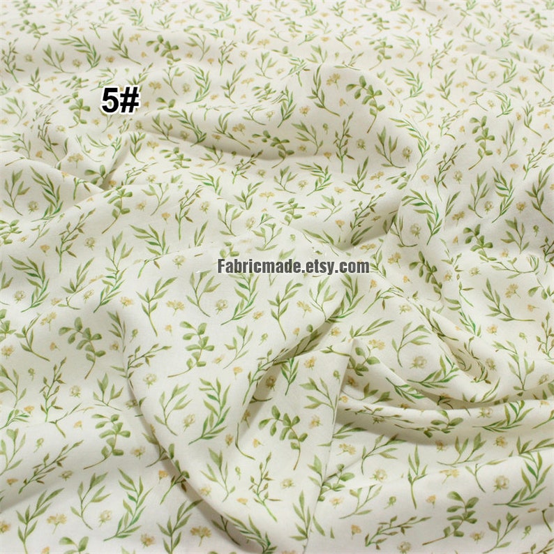 May include: White fabric with a green and yellow floral pattern. The fabric has a subtle texture and a soft, flowing drape. The pattern features small, delicate leaves and flowers. The fabric is ideal for crafting, quilting, and sewing projects.