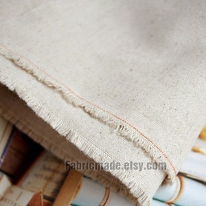 May include: Close-up of a natural off-white linen fabric with a raw edge and a thin orange thread running along the edge. The fabric is folded and the text "Fabricmade.etsy.com" is visible in the bottom right corner.
