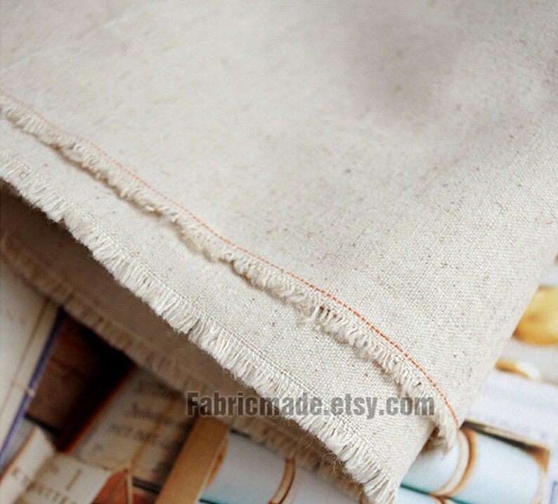 May include: Close-up of a natural off-white linen fabric with a raw edge and a thin orange thread running along the edge. The fabric is folded and the text "Fabricmade.etsy.com" is visible in the bottom right corner.