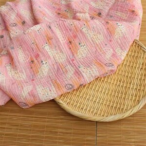 May include: A soft pink muslin fabric featuring a repeating pattern of white cats with colorful polka dots and small black circular scribbles. The lightweight, crinkled textile is draped over a round, woven bamboo tray with a light tan finish.