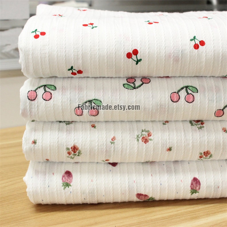 May include: Four white fabric swatches with different patterns. The top swatch has a red cherry pattern, the second has a green cherry pattern, the third has a small pink flower pattern, and the bottom swatch has a pink strawberry pattern. All swatches have a white background and a subtle ribbed texture.