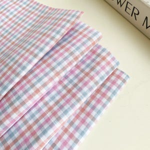 May include: A stack of pastel-colored gingham fabric in shades of pink, blue, and peach. The fabric is neatly folded, showcasing the classic checkered pattern. The fabric appears soft and lightweight, suitable for various craft projects.
