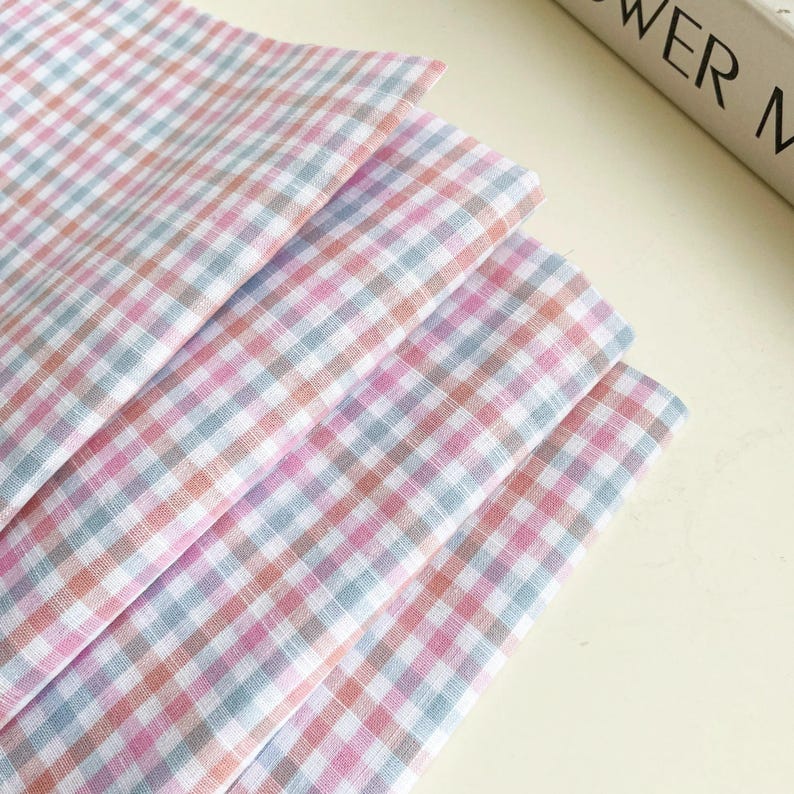 May include: A stack of pastel-colored gingham fabric in shades of pink, blue, and peach. The fabric is neatly folded, showcasing the classic checkered pattern. The fabric appears soft and lightweight, suitable for various craft projects.