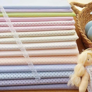 May include: A stack of folded fabric squares in various pastel colors, each featuring a small polka dot pattern. A strip of white lace trim lies diagonally across the fabric stack, and a small doll with blonde yarn hair is visible on the right.