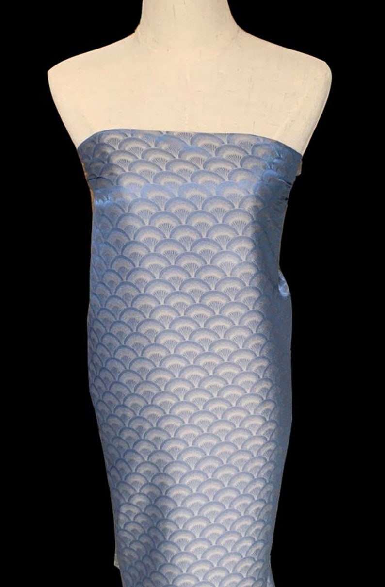 May include: A light blue fabric with a white and light blue patterned design. The pattern is a repeating design of overlapping fan shapes.