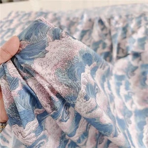 May include: Close-up of a fabric with a wave pattern in shades of blue, white, and pink. The fabric is held up, showing the detailed design of ocean waves. The material appears to be soft and flowing, suitable for clothing or home decor.