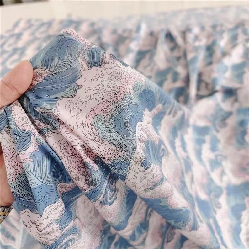 May include: Close-up of a fabric with a wave pattern in shades of blue, white, and pink. The fabric is held up, showing the detailed design of ocean waves. The material appears to be soft and flowing, suitable for clothing or home decor.