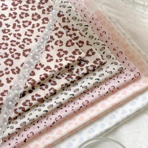 May include: A stack of folded fabric sheets featuring various patterns including brown and black leopard spots, light pink hearts, and pale blue hearts, with a white sheer lace trim featuring small white dots layered over the top edge.
