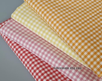 May include: Four stacks of gingham fabric in red, pink, yellow, and orange. Each fabric has a classic checkered pattern. The fabrics are folded and layered, with the website address fabricmade.etsy.com visible on the bottom right.
