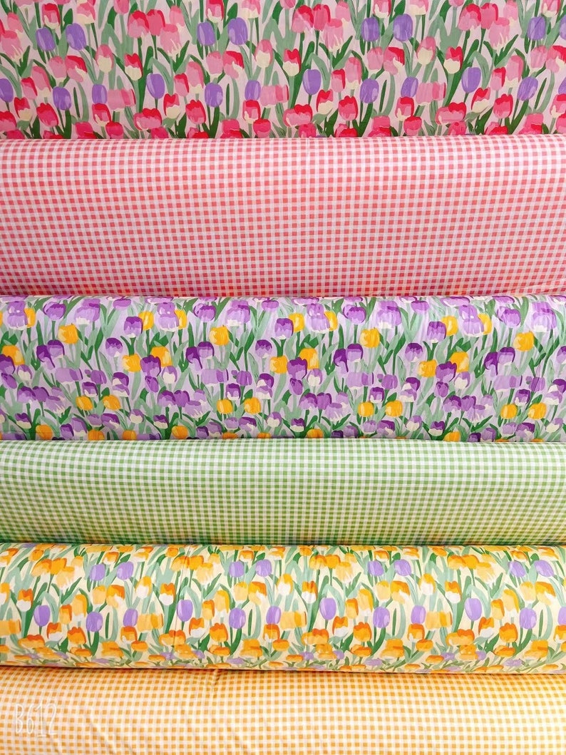 May include: Five rolls of fabric with different patterns. The top roll is pink with white gingham, the second roll is purple with yellow flowers, the third roll is green with white gingham, the fourth roll is yellow with purple flowers, and the bottom roll is orange with white gingham.