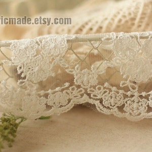 May include: Close-up of delicate white lace trim with a floral pattern. The lace has a scalloped edge and is set against a soft, neutral background. The Etsy shop name fabricmade.etsy.com is visible.