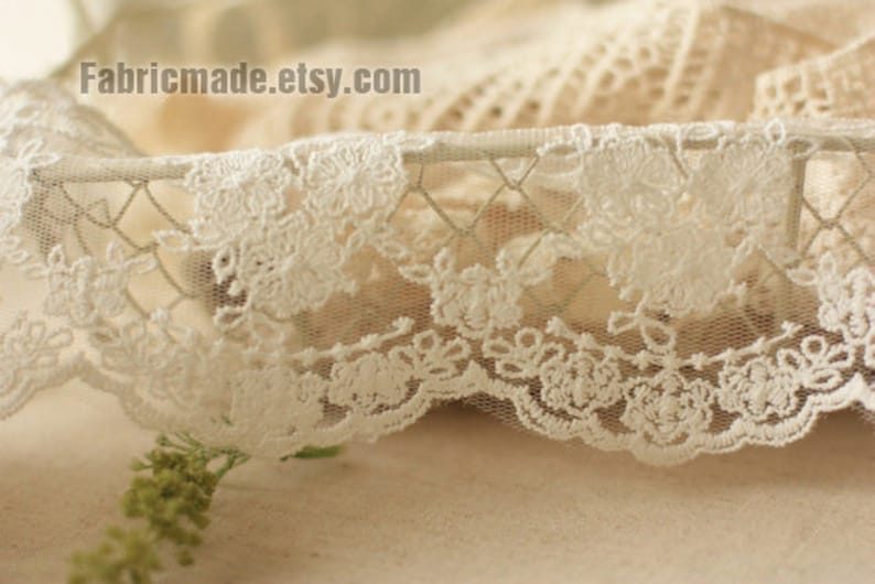 May include: Close-up of delicate white lace trim with a floral pattern. The lace has a scalloped edge and is set against a soft, neutral background. The Etsy shop name fabricmade.etsy.com is visible.