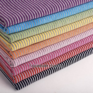 Peut inclure: Une pile de tissu rayé de différentes couleurs, dont le violet, le bleu, le vert, le jaune, l'orange, le rose, le rouge, le noir et le gris. Le tissu est disposé en arc-en-ciel. Le texte "Fabricmade.etsy.com" est visible en bas de la pile.