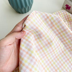 May include: Close-up of a fabric with a gingham pattern in shades of peach, yellow, and white. The fabric is held in a hand, with the edges of the fabric visible. The fabric appears to be soft and lightweight.