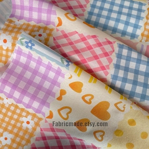 Könnte beinhalten: Nahaufnahme von farbenfrohen Stoffrollen mit verschiedenen Mustern. Die Muster umfassen Vichy-Karos in Rosa, Lila und Blau sowie Blumen- und Herzmotive in Orange und Gelb. Der Stoff ist wahrscheinlich Baumwolle oder ein ähnliches Material.