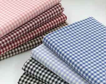 May include: Stacks of gingham fabric in various colors, including pink, burgundy, brown, light blue, dark blue, and black. The fabric is neatly folded, showcasing the classic checkered pattern. Ideal for sewing and crafting projects.