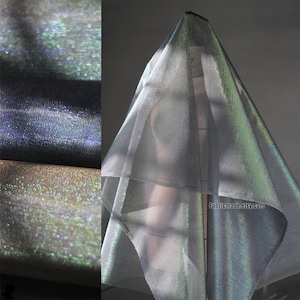 May include: Iridescent fabric in shades of silver, blue, and gold. The fabric is draped over a form, showcasing its shimmering qualities. The image also shows rolls of the fabric, highlighting its texture and color variations. The website address fabricmade.etsy.com is visible.