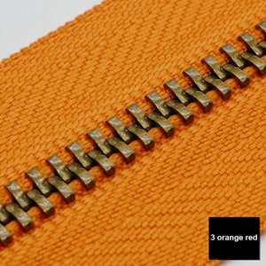 May include: Close-up of an orange zipper tape with a brass zipper chain. The zipper chain has a hammered finish. The text "3 orange red" is visible in the lower right corner of the image.