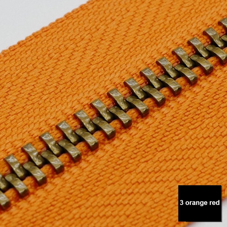 May include: Close-up of an orange zipper tape with a brass zipper chain. The zipper chain has a hammered finish. The text "3 orange red" is visible in the lower right corner of the image.