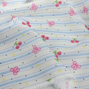 May include: White fabric patterned with blue stripes, pink bows, and red cherries. The fabric also features small black dots and yellow star shapes. The design is repeated across the fabric, creating a whimsical pattern.