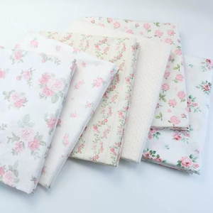 May include: Several folded fabric pieces with floral designs. The fabrics showcase a variety of patterns, including pink roses on white, dotted patterns, and vertical stripes with floral motifs. The overall style is soft and feminine, suitable for crafting.
