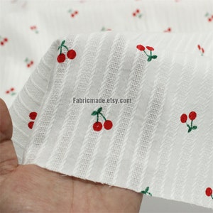 May include: White cotton fabric with red cherries and thin white stripes. The fabric has a textured surface. "Fabricmade.etsy.com" is printed on the fabric.